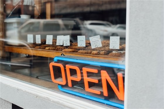 A neon open sign sits in front of a tray of cookies in a bakery's window